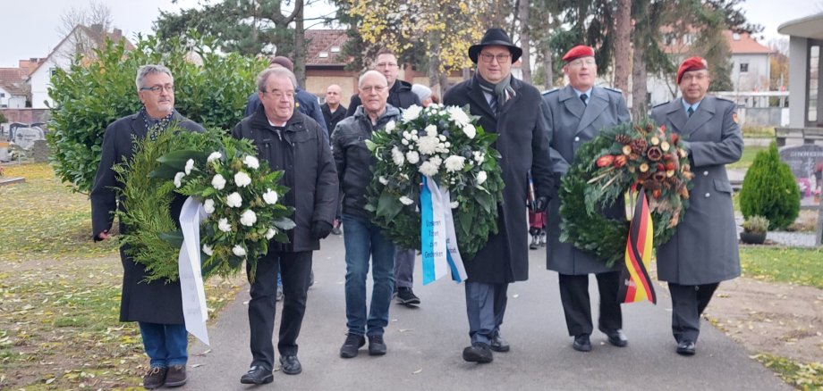 Männer auf einem Friedhofsweg, die drei Trauerkränze tragen