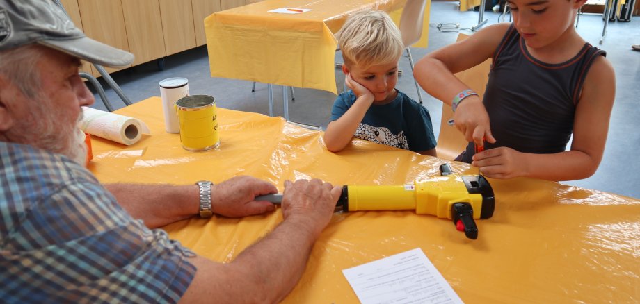 Ein älterer Mann repariert eine Maschine. 2 Kinder schauen zu.